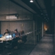 A dark image of a glass-walled office with people meeting with laptops at a table. Featured in an article on toxic cybersecurity culture.
